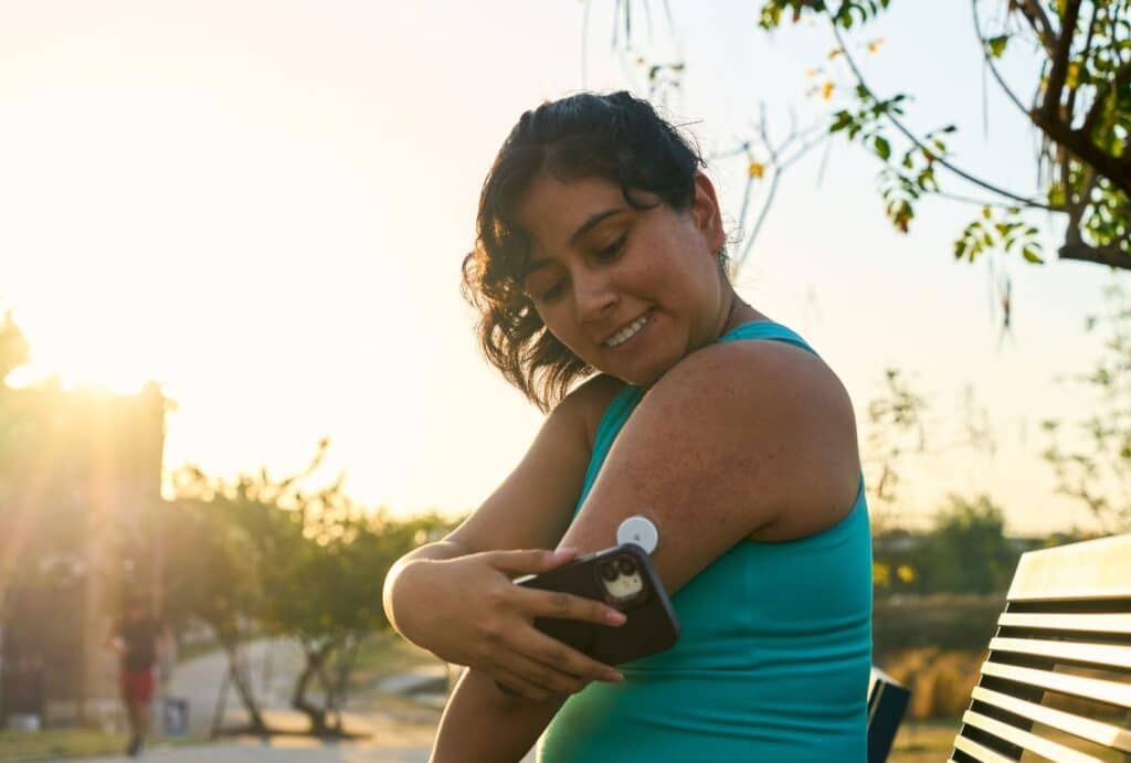 אישה צעירה עם סוכרת בודקת רמת סוכר באמצעות חיישן CGM על הזרוע באמצעות הסמארטפון בפארק בשקיעה